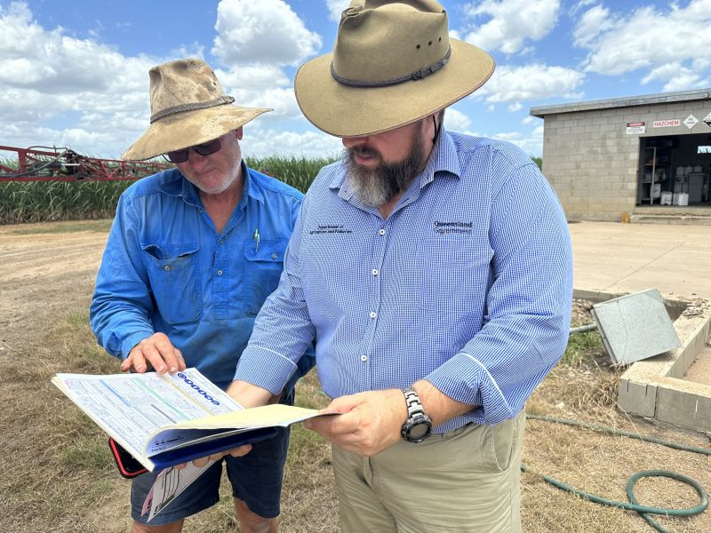 2 people wearing hats and blue shirts looking at a book. Farm in background
