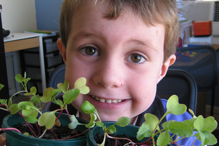 Primary school student holding 2 pot plants below his face and looking at camera.