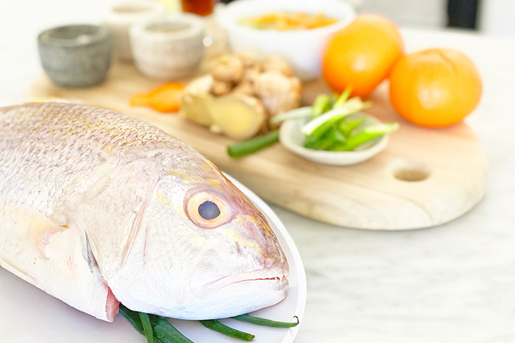 Filleted fish with head on chopping board surrounded by food items in preparation for cooking.