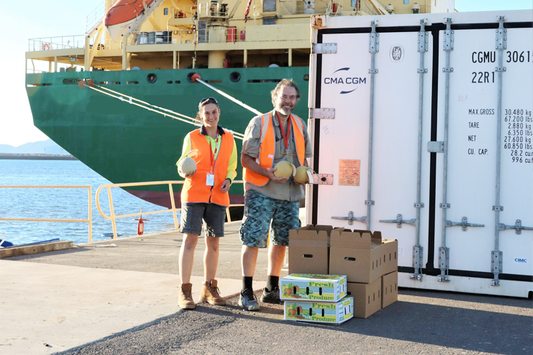 Two people holding fruit and standing on a jetty in front of a shipping container and boat.