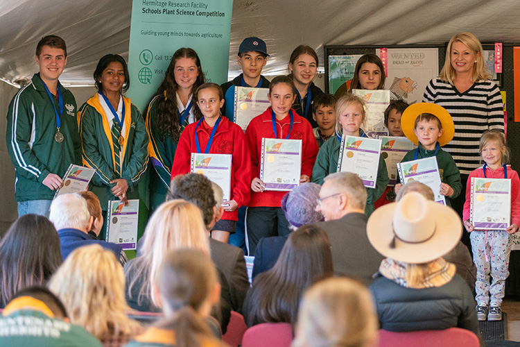 Group of school children (aged primary to high school) standing on stag with Ranger Stacey in front of seated audience, showing their award certificates.